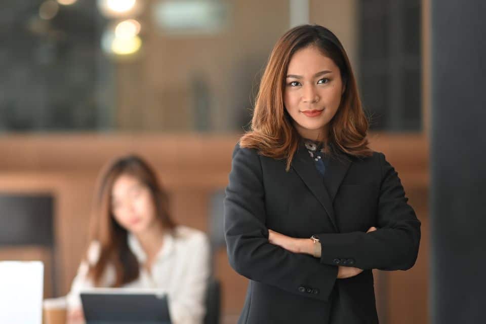 Confident female leader standing with arms crossed in a professional setting, representing the shift from inner critic to inner champion discussed in the article.